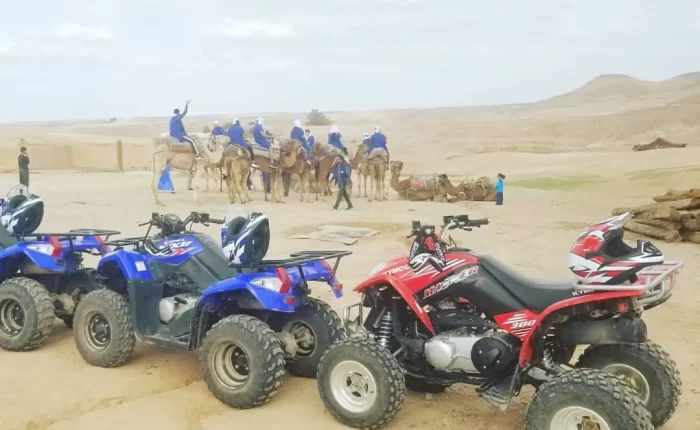 A group of adventurers partaking in a camel ride across the undulating dunes of the Agafay Desert, with the Atlas Mountains faintly visible in the distance. In the foreground, parked quad bikes and buggies hint at the action-packed ride to follow