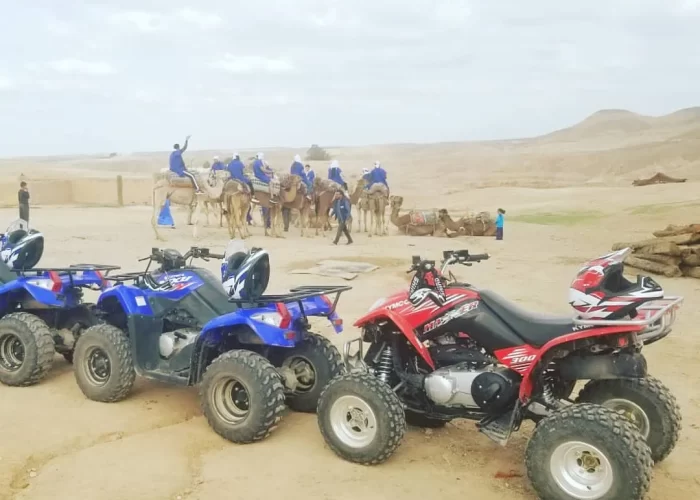 A group of adventurers partaking in a camel ride across the undulating dunes of the Agafay Desert, with the Atlas Mountains faintly visible in the distance. In the foreground, parked quad bikes and buggies hint at the action-packed ride to follow