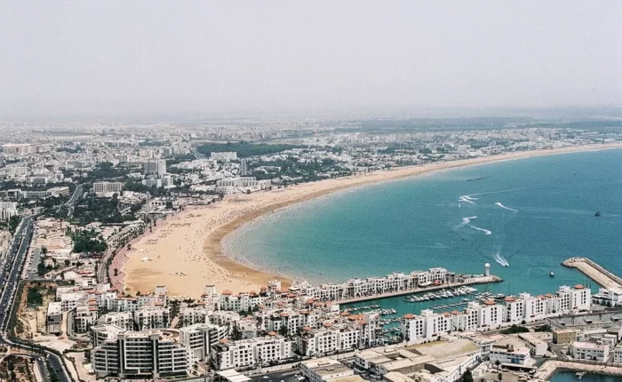 Bright sunshine illuminates the modern coastal cityscape of Agadir, Morocco, as travelers from a full day trip from Marrakech to Agadir City stroll leisurely along the vibrant beach promenade,