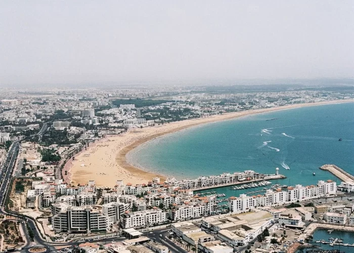 Bright sunshine illuminates the modern coastal cityscape of Agadir, Morocco, as travelers from a full day trip from Marrakech to Agadir City stroll leisurely along the vibrant beach promenade,