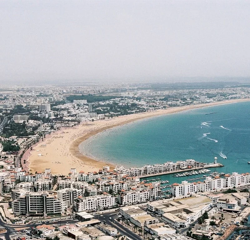 Bright sunshine illuminates the modern coastal cityscape of Agadir, Morocco, as travelers from a full day trip from Marrakech to Agadir City stroll leisurely along the vibrant beach promenade,